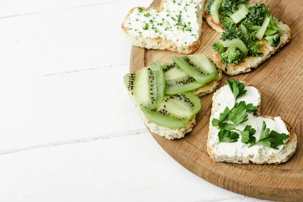 Heart shaped canape with creamy cheese, broccoli, microgreen, parsley and kiwi on board on white wooden surface — Stock Photo