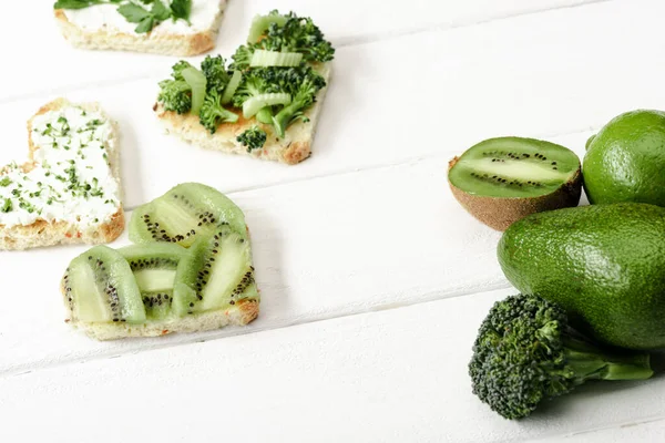 Heart shaped canape with creamy cheese, broccoli, microgreen, parsley and kiwi near green fruits and vegetables on white wooden surface — Stock Photo