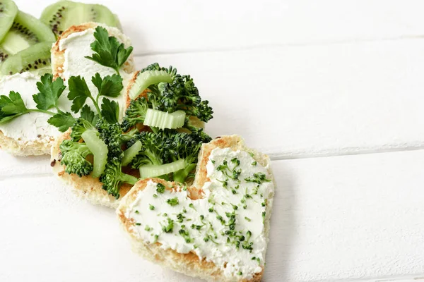 Tartina a forma di cuore con formaggio cremoso, broccoli, microverde, prezzemolo e kiwi su superficie di legno bianca — Foto stock