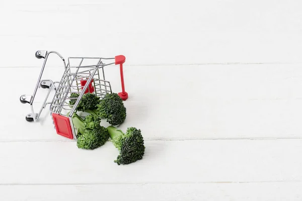 Fresh green broccoli scattered from shopping cart on white wooden surface — Stock Photo