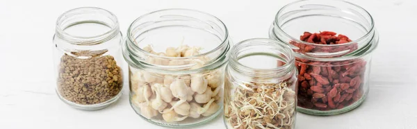 Bayas y brotes de goji en frascos de vidrio sobre superficie de madera blanca, orientación panorámica — Stock Photo