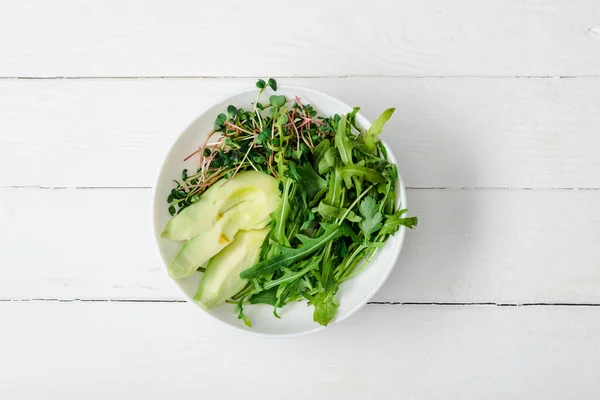 Top view of arugula, avocado and microgreen in bowl on white wooden surface — Stock Photo