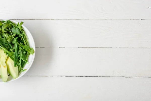 Vue du dessus de la roquette, avocat dans un bol sur une surface en bois blanc — Photo de stock