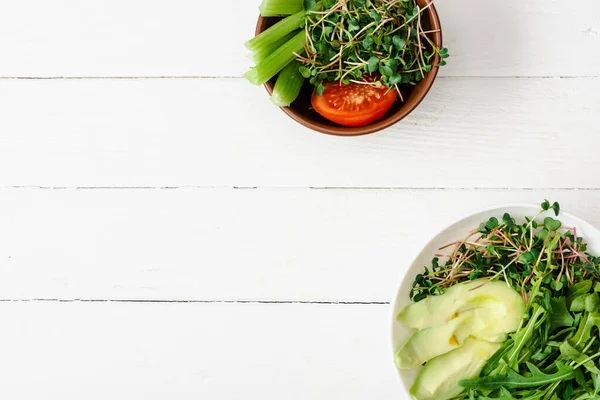 Top view of fresh vegetables with avocado and microgreen in bowls on white wooden surface — Stock Photo