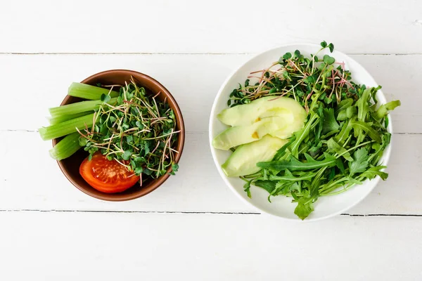 Вид сверху на свежие овощи с авокадо и микрозеленый в мисках на белой деревянной поверхности — стоковое фото