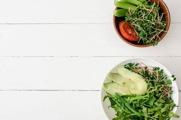 Top view of fresh vegetables with avocado and microgreen in bowls on white wooden surface — Stock Photo