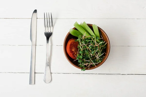 Vista superior de tomate, apio y microgreen en tazón sobre superficie de madera blanca con cubiertos - foto de stock