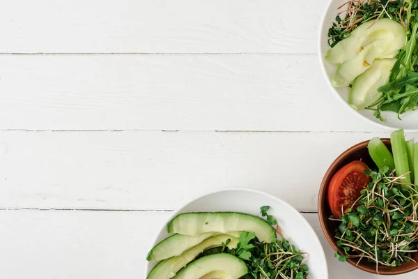 Вид зверху на свіжі овочі з авокадо і мікрозеленим в мисках на білій дерев'яній поверхні — стокове фото