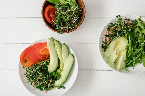 Draufsicht auf frisches Gemüse mit Avocado und Microgreen in Schalen auf weißer Holzoberfläche — Stockfoto