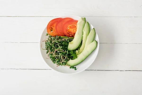 Vista superior de tomate, aguacate y microgreen en tazón sobre superficie de madera blanca - foto de stock