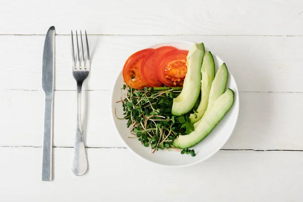 Top view of tomato, avocado and microgreen in bowl on white wooden surface with cutlery — Stock Photo