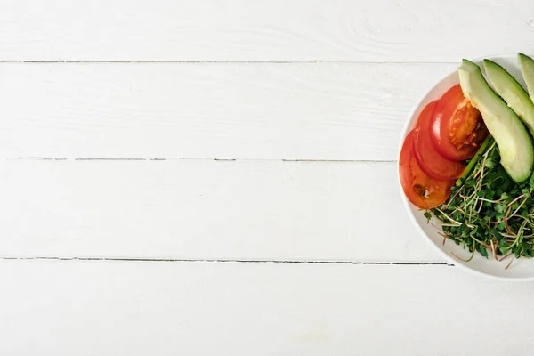 Vista superior de tomate, aguacate y microgreen en tazón sobre superficie de madera blanca - foto de stock