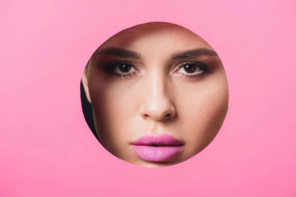 Женщина с дымчатыми глазами и розовыми губами, смотрящая в камеру через отверстие в бумаге — стоковое фото
