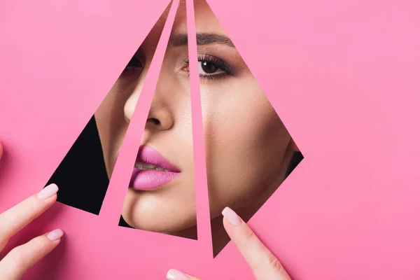 Belle femme aux yeux fumés et aux lèvres roses regardant la caméra à travers des trous triangulaires et touchant du papier sur du noir — Photo de stock