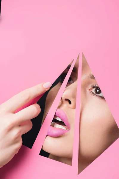 Woman with open mouth and pink lips looking across triangular holes and touching paper with hand on black background — Stock Photo