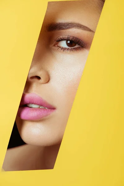 Женщина с макияжем смотрит в камеру через четырехугольное отверстие в желтой бумаге — стоковое фото