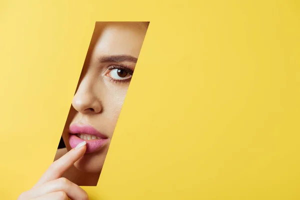 Woman looking at camera across quadrangular hole in yellow paper and touching pink lip — Stock Photo