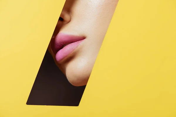 Partial view of female face with pink lips in quadrilateral hole in yellow paper on black — Stock Photo