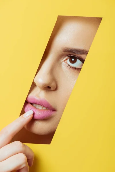 Woman looking across quadrangular hole in yellow paper and touching pink lip — Stock Photo