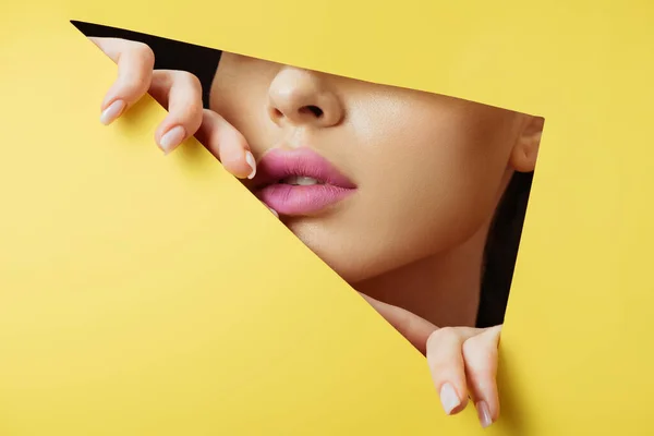 Cropped view of woman with pink lips touching yellow paper across triangular hole on black background — Stock Photo