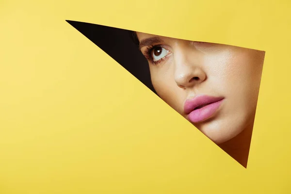 Woman with pink lips looking across triangular hole in yellow paper on black background — Stock Photo