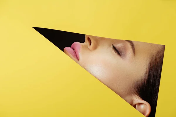 Female face with with pink lips and closed eyes in triangular hole in yellow paper on black — Stock Photo