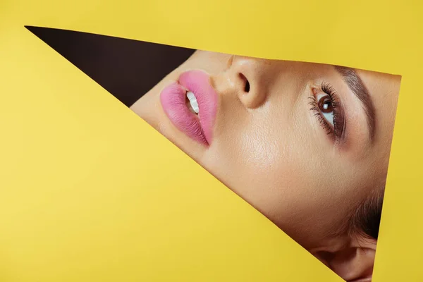 Girl with pink lips looking across triangular hole in yellow paper on black — Stock Photo