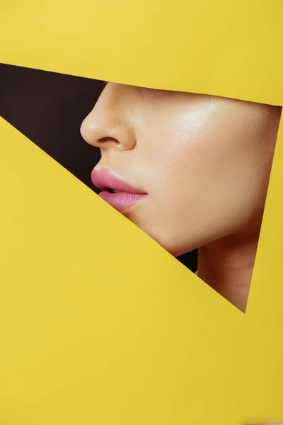 Vista cortada da face feminina com lábios cor-de-rosa em furo triangular em papel amarelo sobre preto — Fotografia de Stock