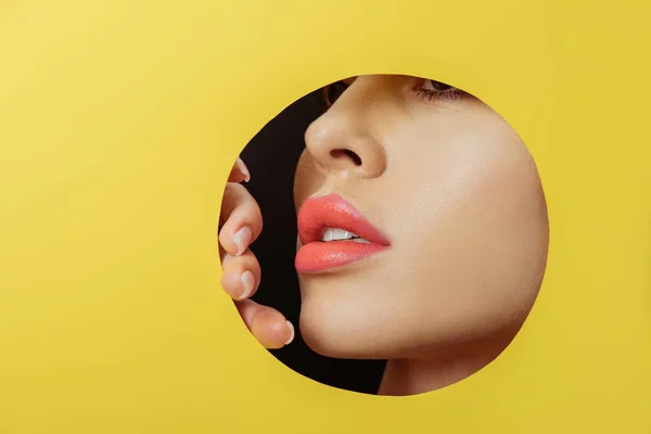 Cara femenina con labios y dedos de coral en agujero redondo en papel amarillo sobre negro - foto de stock