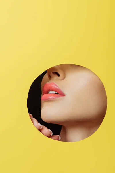 Vue recadrée du visage féminin avec lèvres corail et la main dans un trou rond en papier jaune sur noir — Photo de stock