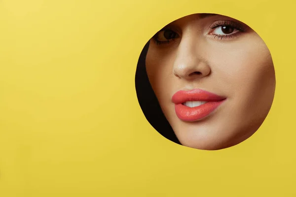Woman with coral lips and smoky eyes looking at camera and smiling across round hole in yellow paper on black — Stock Photo