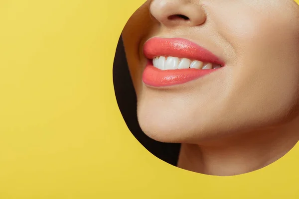 Vista recortada de mujer con labios de coral sonriendo en agujero redondo en papel amarillo sobre negro - foto de stock