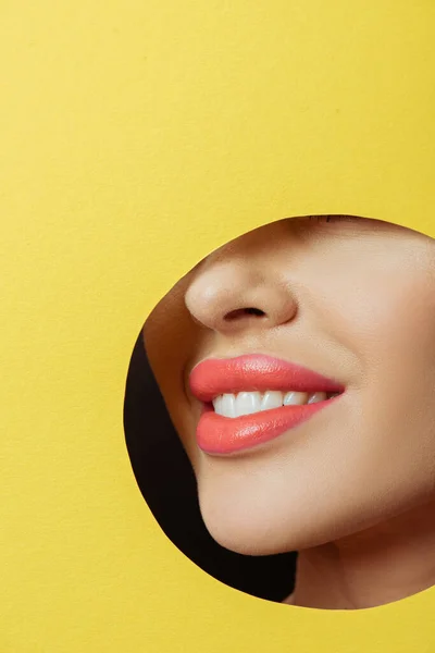 Cropped view of woman with coral lips smiling in round hole in yellow paper on black background — Stock Photo