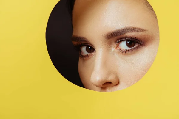 Girl with smoky eyes looking at camera across hole in yellow paper on black background — Stock Photo