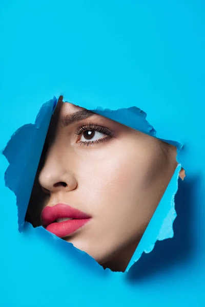 Красивая женщина с розовыми губами, смотрящая через разорванную голубую бумагу — стоковое фото