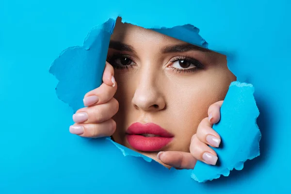 Beautiful woman with smoky eyes and pink lips looking at camera across hole and touching blue paper — Stock Photo