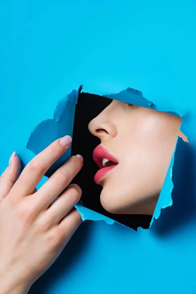 Femme aux lèvres roses et bouche ouverte dans un trou touchant du papier bleu sur fond noir — Photo de stock