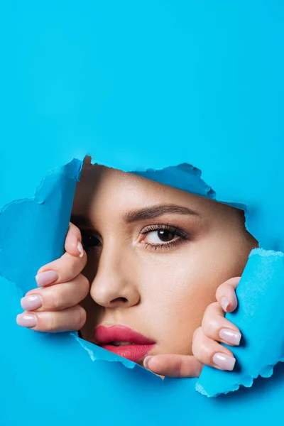 Beautiful woman with pink lips looking at camera across hole and touching bright blue paper — Stock Photo