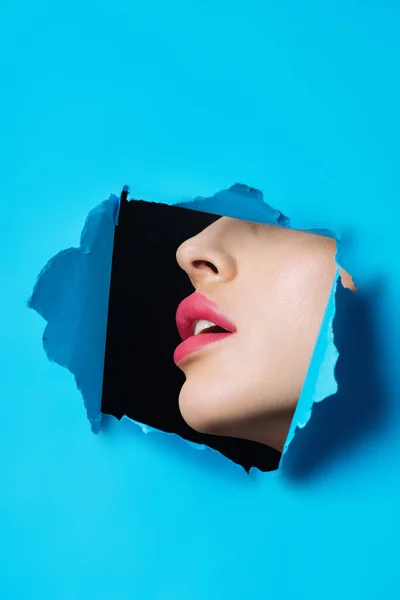 Female face with pink lips in hole in blue paper on black — Stock Photo