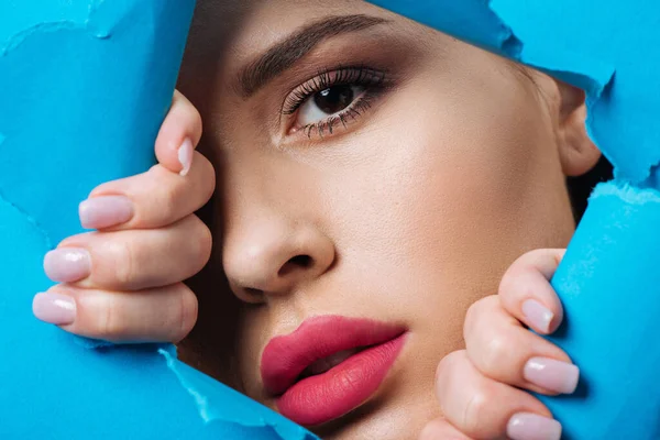 Attraktive Frau mit rosafarbenen Lippen und rauchigen Augen, die in die Kamera blickt und hellblaues Papier berührt — Stockfoto