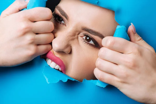 Woman with pink lips and smoky eyes touching and biting ripped blue paper across hole — Stock Photo