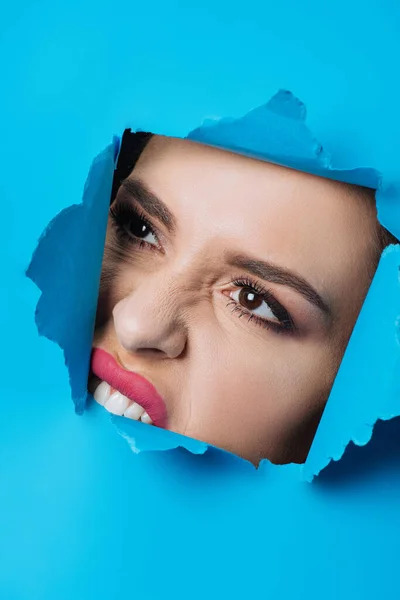 Женщина с розовыми губами и дымчатыми глазами, кусающими рваную голубую бумагу через отверстие — стоковое фото