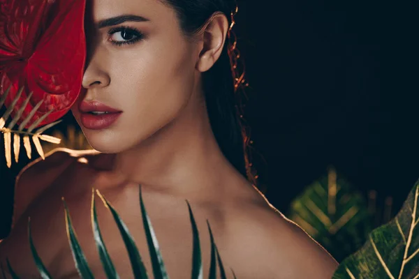 Selective focus of sexy woman near leaves covering face with red anthurium and looking at camera isolated on black — Stock Photo