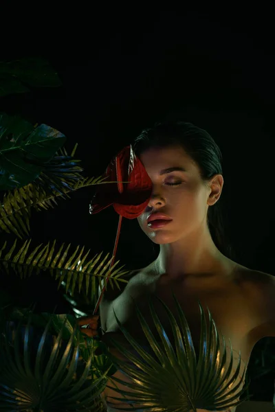 Mujer con los ojos cerrados cubriendo la cara con flores cerca de hojas aisladas en negro - foto de stock