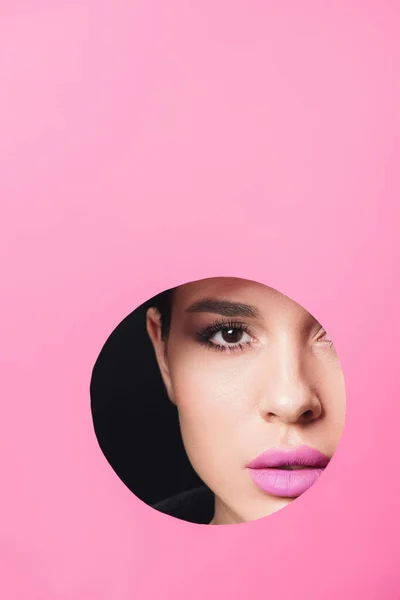 Beautiful girl with smoky eyes and pink lips looking at camera across hole in paper on black — Stock Photo