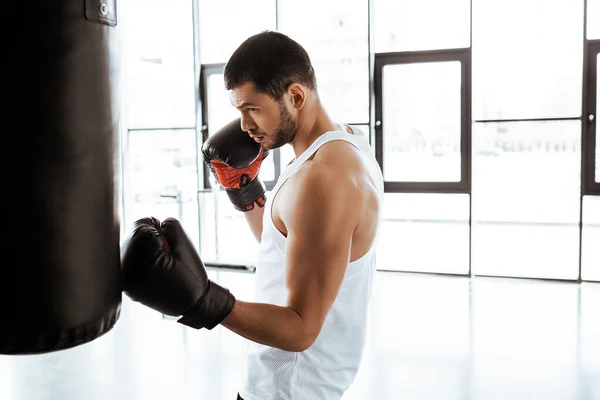 Visão lateral de belo desportista no treinamento de luvas de boxe com saco de perfuração no centro esportivo — Fotografia de Stock