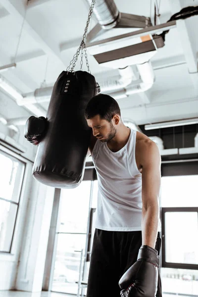 Измученный спортсмен в боксёрских перчатках стоит рядом с боксёрской грушей — стоковое фото