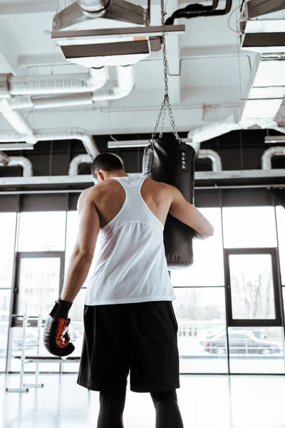 Visão traseira do desportista em luvas de boxe tocando saco de perfuração enquanto em pé no centro de esportes — Fotografia de Stock
