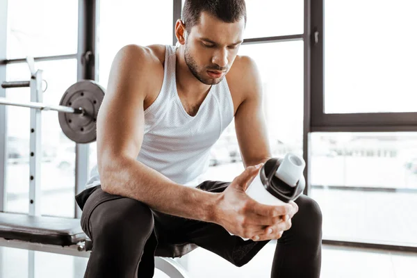 Deportista cansado sosteniendo botella deportiva con batido de proteínas en el gimnasio — Stock Photo