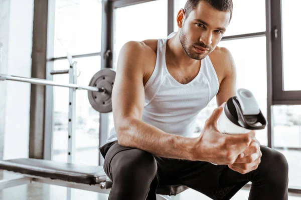 Apuesto deportista sosteniendo botella deportiva con batido de proteínas y mirando a la cámara en el gimnasio - foto de stock